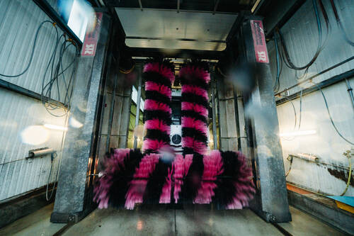 Ein LKW fährt durch eine Waschanlage. Die pink-schwarzen Bürsten reinigen gerade die Front und verdecken den LKW teilweise.