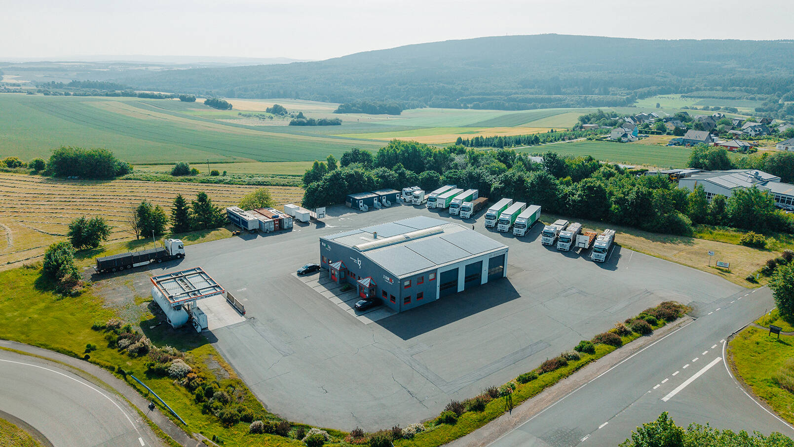 Drohnenaufnahme des Firmengeländes in Hochscheid: Mehrere LKWs stehen auf dem Hof, zentral befindet sich das Firmengebäude, am linken Bildrand ist eine Tankstelle zu sehen.